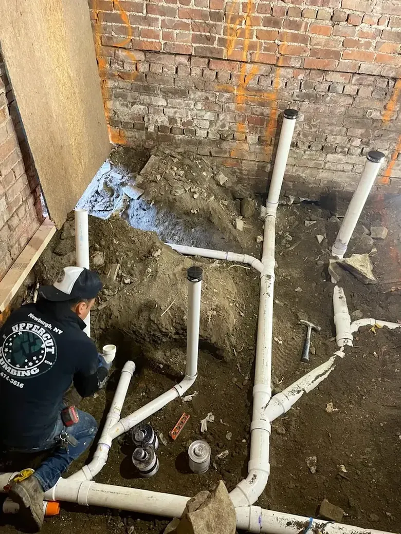 An Uppercut Plumbing technician in a branded sweatshirt setting new PVC drain lines in a basement